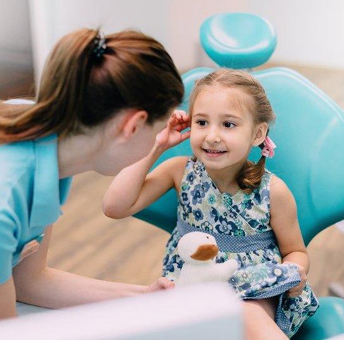Little girl sitting in dental examination chair and playing with her ear while talking to a dental assistant 