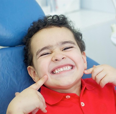 Young boy sitting in dental chair smiling and pointing to his teeth
